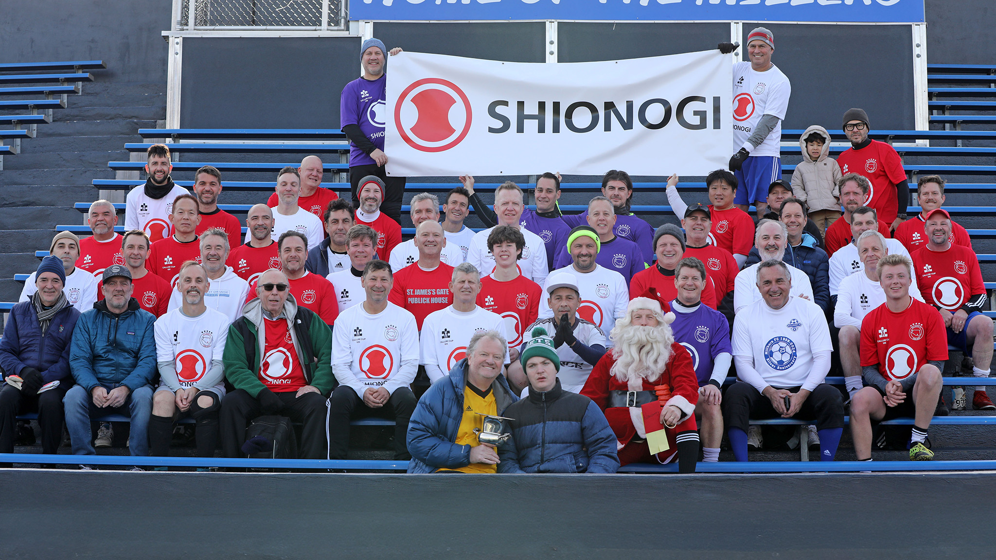 Group photo of Callum Cup players seated in the Millburn High School bleachers, wearing red, white, and purple jerseys, with a Shionogi banner displayed behind them.