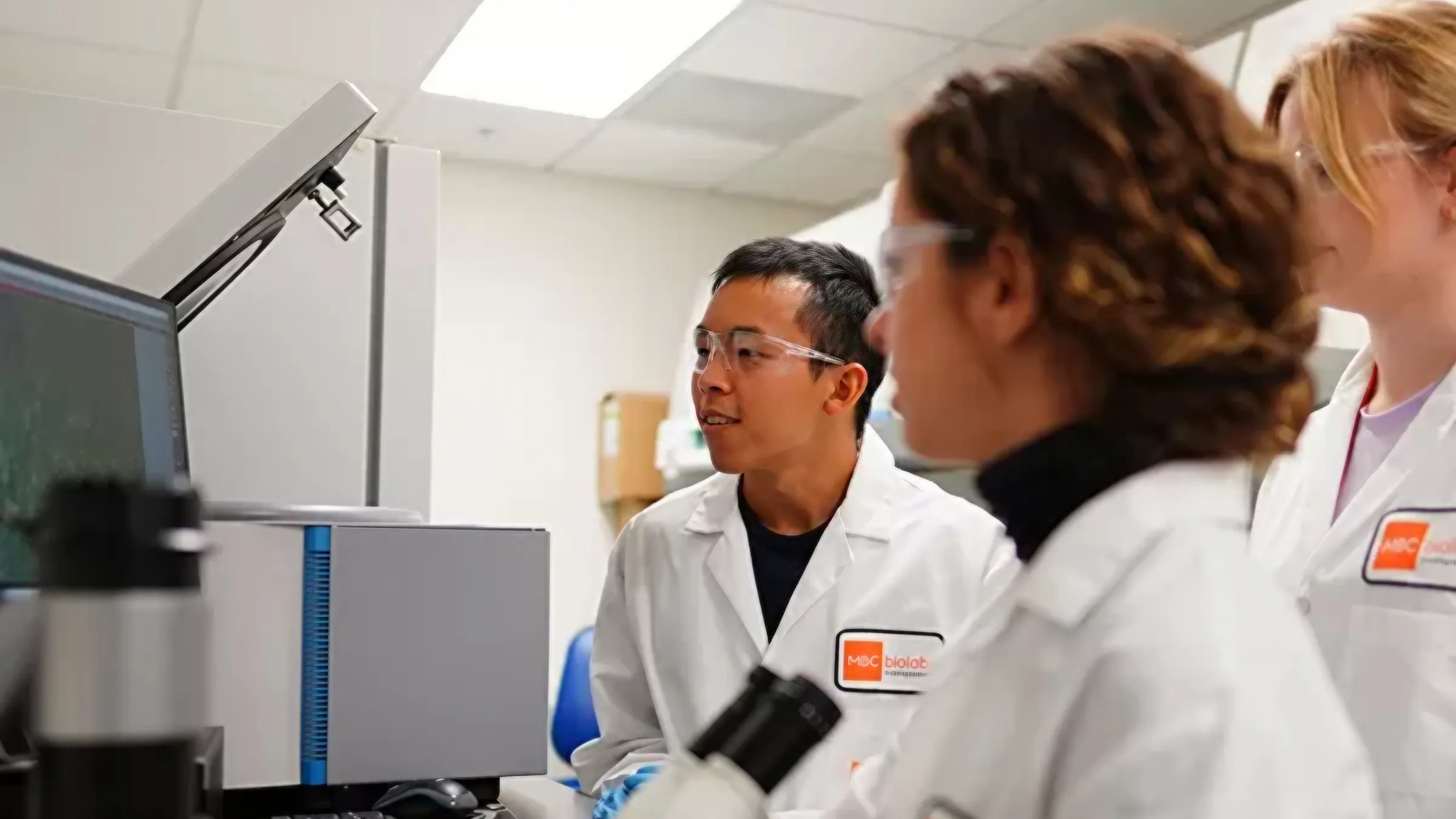 Researchers in white lab coats reviewing a computer display beside a microscope.