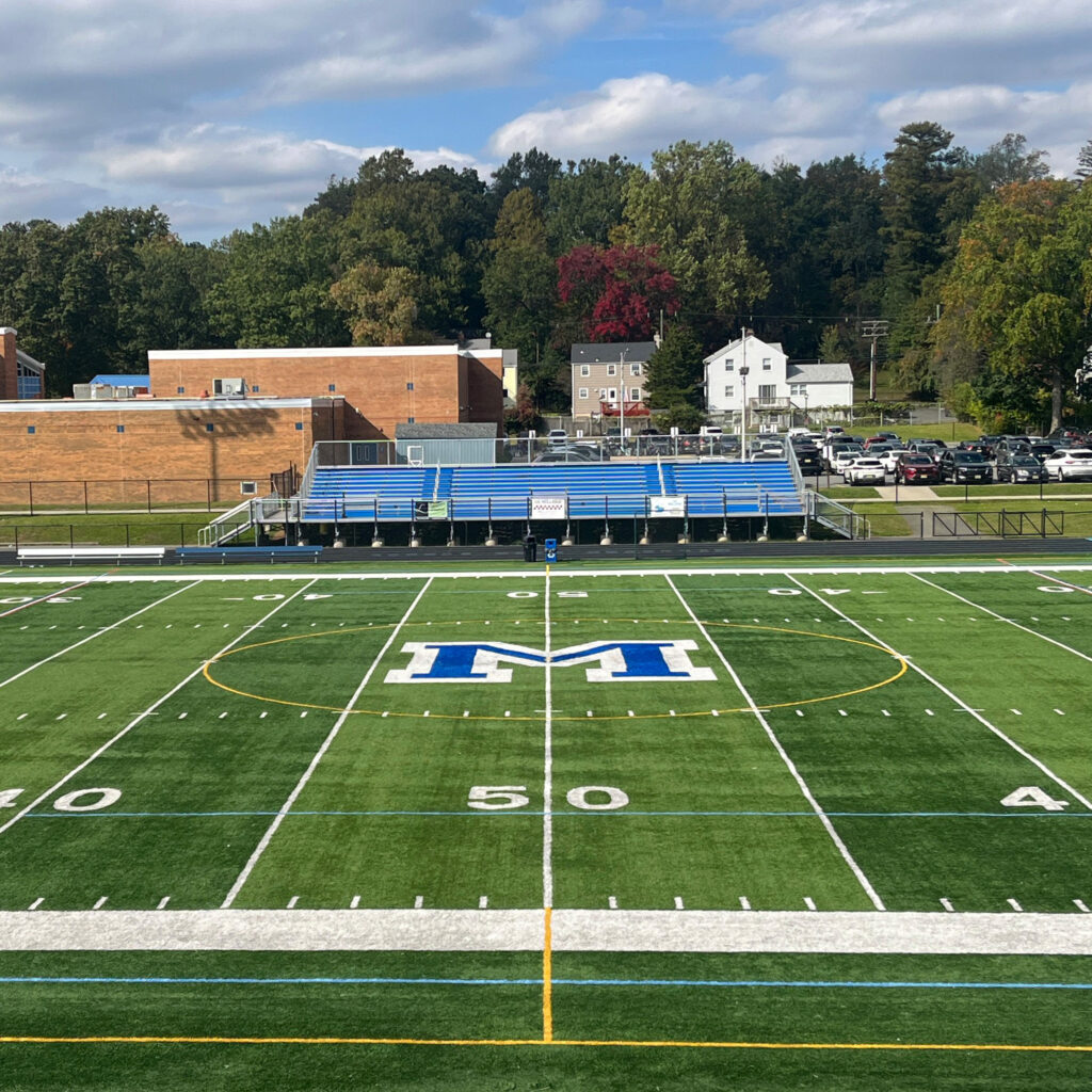 Wide view of the Millburn High School athletic field showing the 50-yard line, blue stadium bleachers, and school buildings with trees in the background.