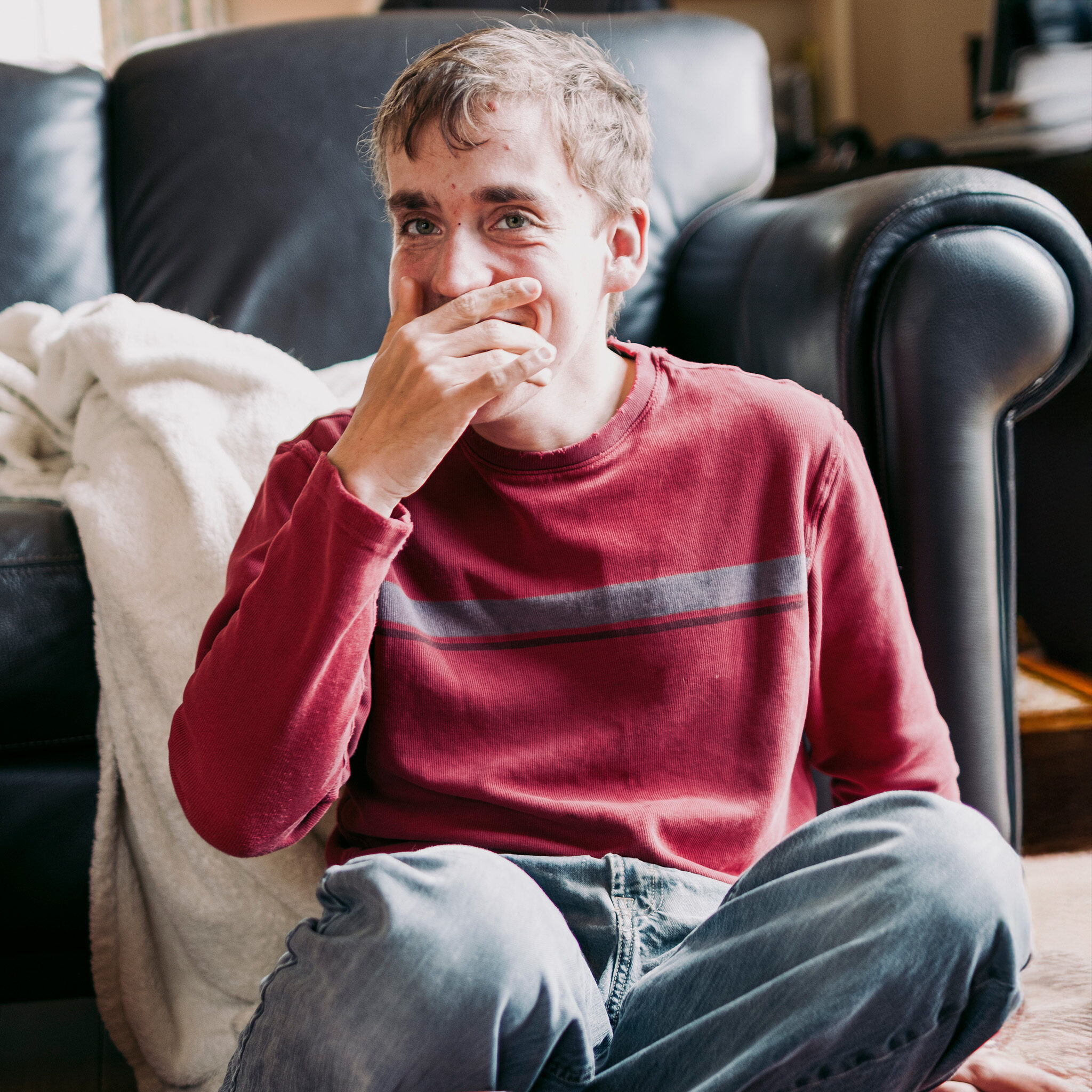 A young man with Fragile X syndrome smiling while sitting on the floor at home.